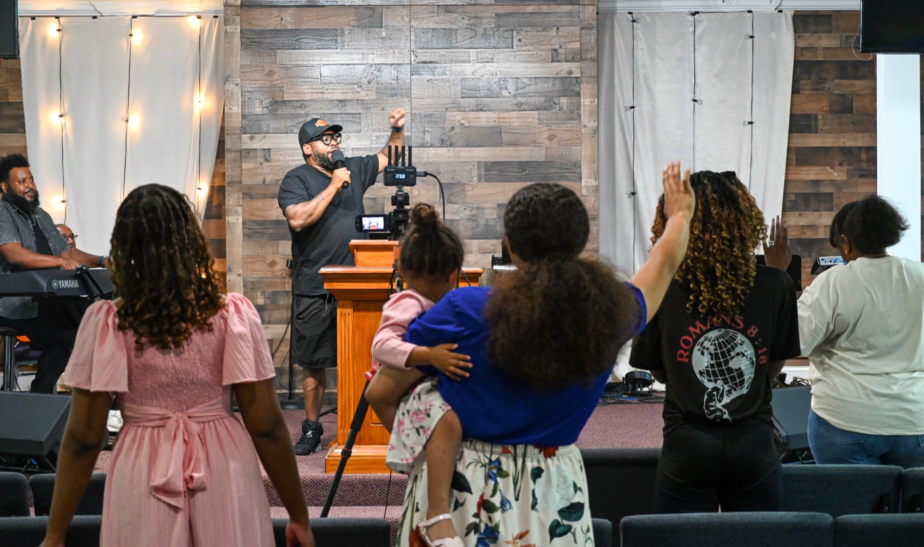 The congregation standing with hands raised as the pastor speaks from the podium, a keyboard player to one side.